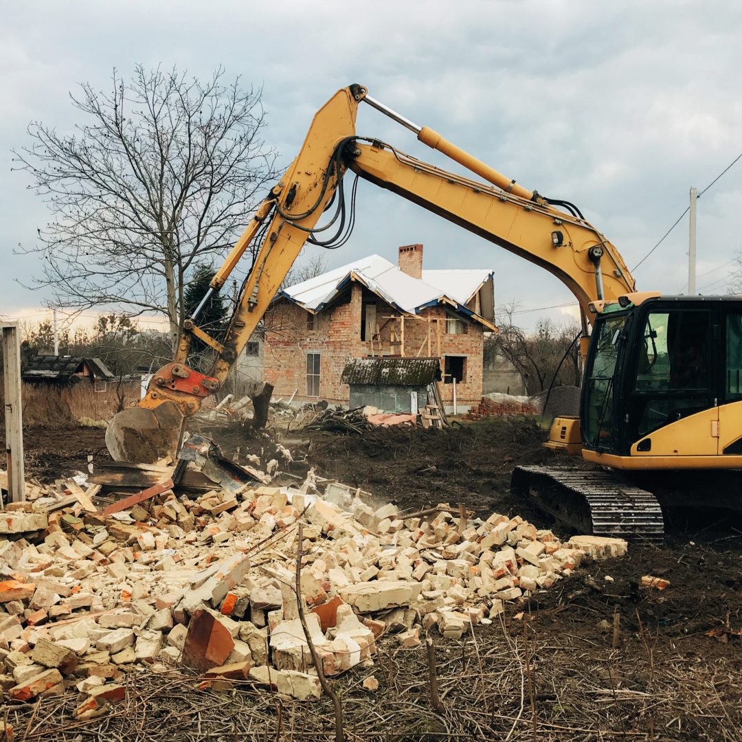 Excavator destroying brick house on land in countryside. Bulldozer clearing land from old bricks and concrete from walls with dirt and trash. Backhoe machinery ruining house